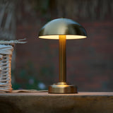Endon Ashfield LED table lamp in brass with domed shade and fluted stem, glowing warmly on a wooden ledge beside a woven basket outdoors.
