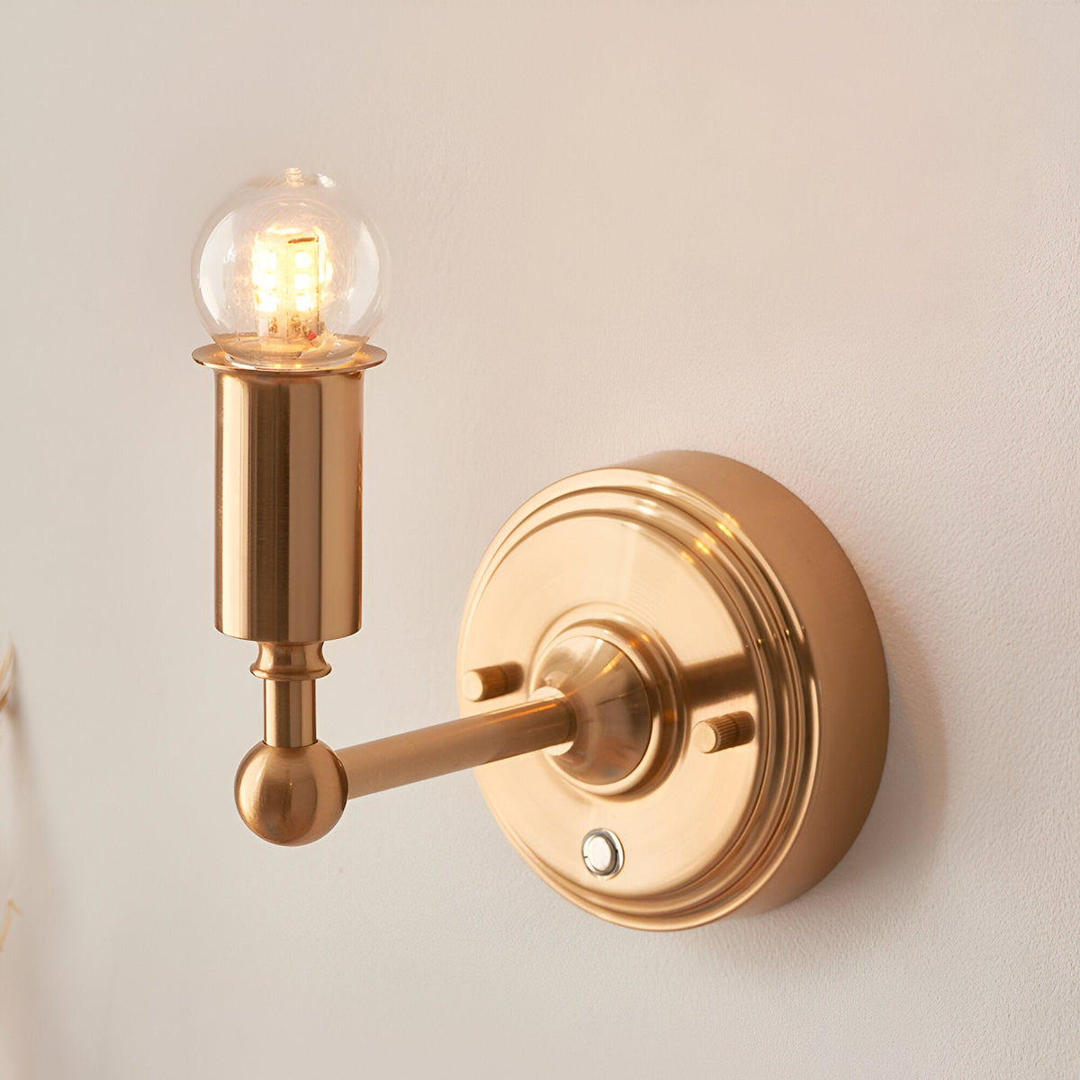 Endon Ashstead LED rechargeable wall light in brushed aged brass with a small clear globe bulb and round backplate, mounted on a pale wall.