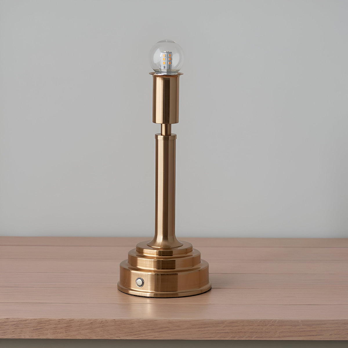 Danebourne LED Table Lamp in brushed aged brass with a stepped base, slim column, exposed globe bulb and touch dimmer, shown on a wooden surface.