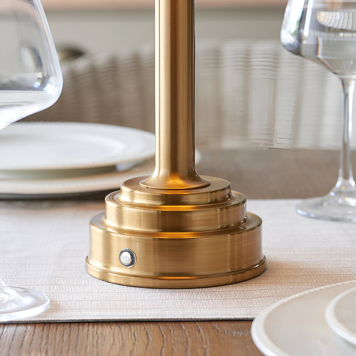 Danebourne LED Table Lamp base in brushed aged brass with stepped tiers and touch dimmer button, shown on a dining table beside glassware and plates.