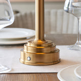 Danebourne LED Table Lamp base in brushed aged brass with stepped tiers and touch dimmer button, shown on a dining table beside glassware and plates.