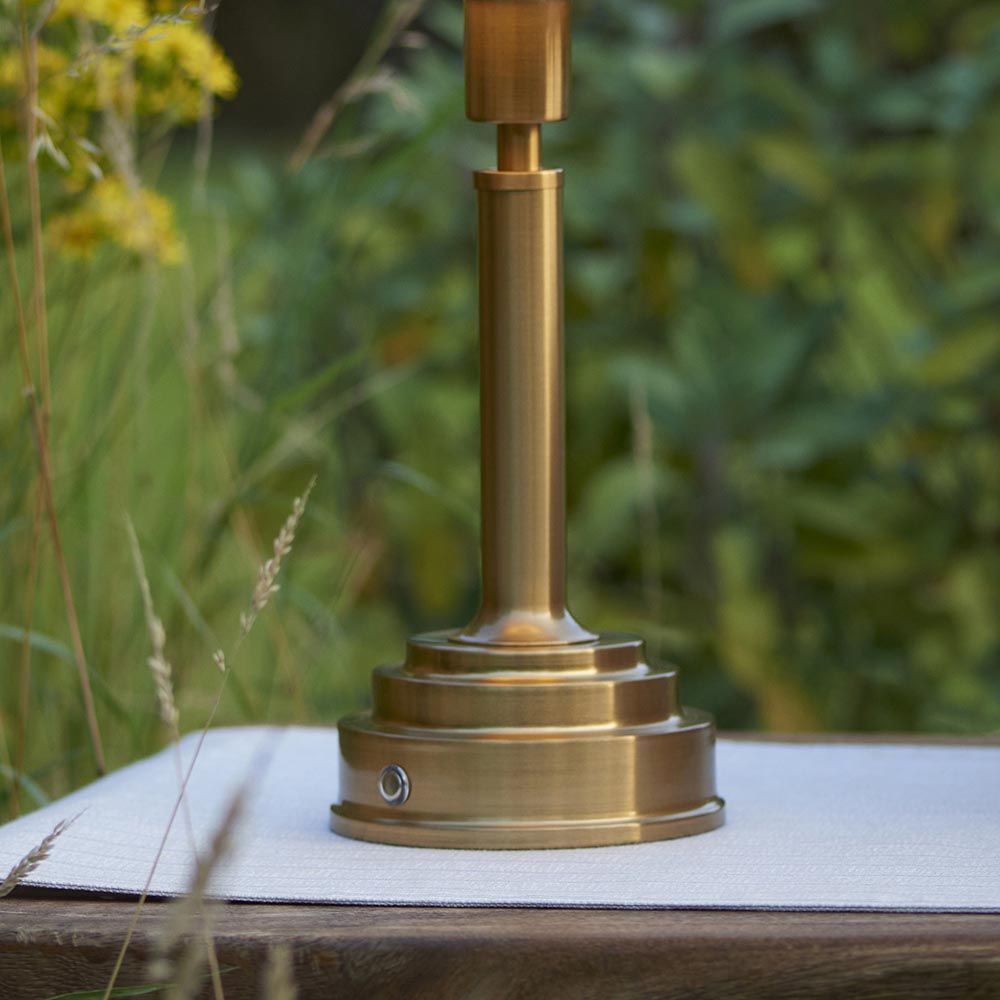 Danebourne LED Table Lamp base in brushed antique brass with stepped tiered design and touch dimmer button, shown outdoors on a table.