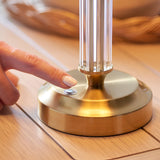 Finger pressing the touch dimmer button on the brushed brass base of the Endon Carlwood LED table lamp, with clear fluted acrylic column above.