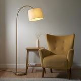 Endon Bevbourne arched floor lamp in brass with tilting natural fabric drum shade, beside a mustard velvet armchair and wooden side table.