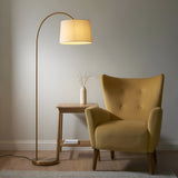 Endon Bevbourne arched floor lamp in brass with tilting natural fabric drum shade, beside a mustard velvet armchair and wooden side table.