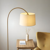 Endon Bevbourne arched floor lamp in brass with tilting natural fabric drum shade, beside a wooden side table and yellow armchair.