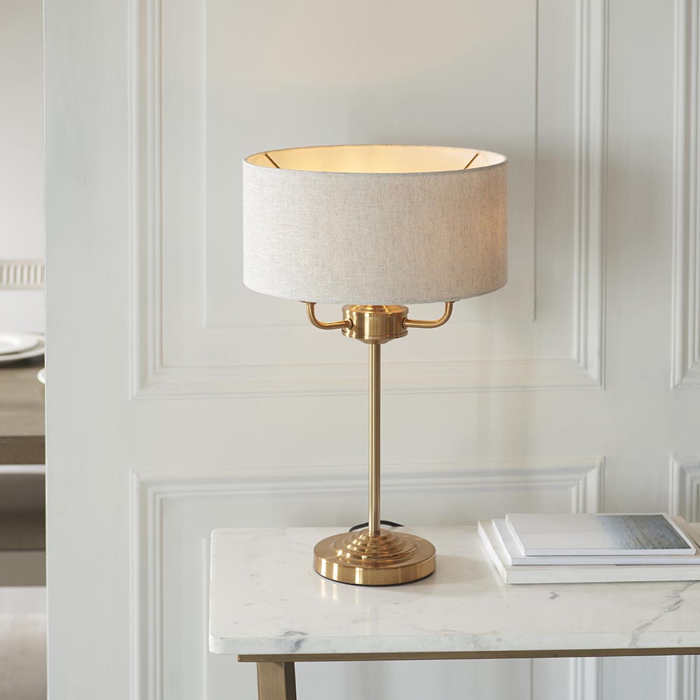 Brenley 3 Light Table Lamp in brass, with a stepped round base, slender stem, and linen drum shade, shown lit on a marble console table.