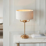 Brenley 3 Light Table Lamp in brass, with a stepped round base, slender stem, and linen drum shade, shown lit on a marble console table.