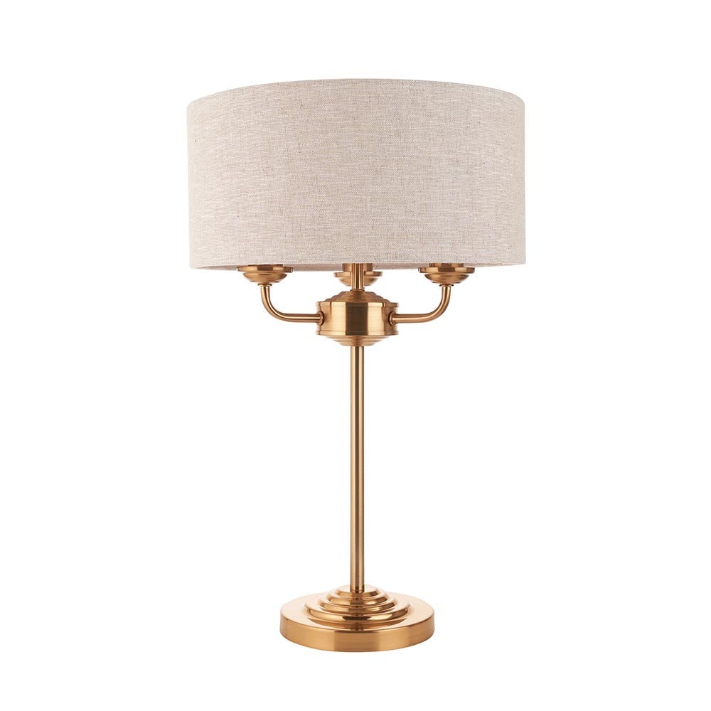 Brenley 3 Light Table Lamp in brass, with stepped round base, tall stem, three-armed fitting and a beige linen drum shade, shown on a plain white background.