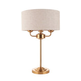 Brenley 3 Light Table Lamp in brass, with stepped round base, tall stem, three-armed fitting and a beige linen drum shade, shown on a plain white background.