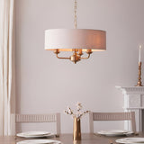 Brenley 3 Light Pendant in vintage brass with a natural linen drum shade, hung above a dining table set with white plates and a brass vase.
