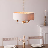 Brenley 3 Light Pendant in vintage brass with a natural linen drum shade, hung on chain above a dining table in a light-toned room.
