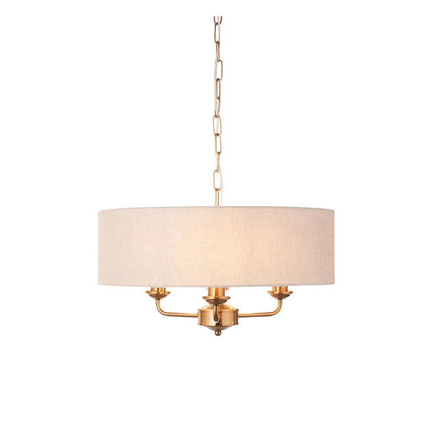 Brenley 3 Light Pendant in vintage brass with a natural linen drum shade, three exposed candle-style arms, and hanging chain, shown on a plain white background.