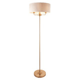 Endon Brenley 3 light floor lamp in vintage brass with a wide natural linen drum shade and slim central stem on a round base, shown on a white background.