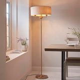 Endon Brenley 3 light floor lamp in brass with a wide natural linen drum shade and triple-arm fitting, set beside a dining table and window.