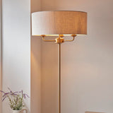 Endon Brenley 3 Light Floor Lamp in brass with a wide natural linen drum shade, shown lit in a corner beside lavender stems.