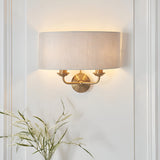 Endon Brenley 2 light wall light in vintage brass with a natural linen drum shade, mounted on a white panelled wall above a vase of dried grasses.