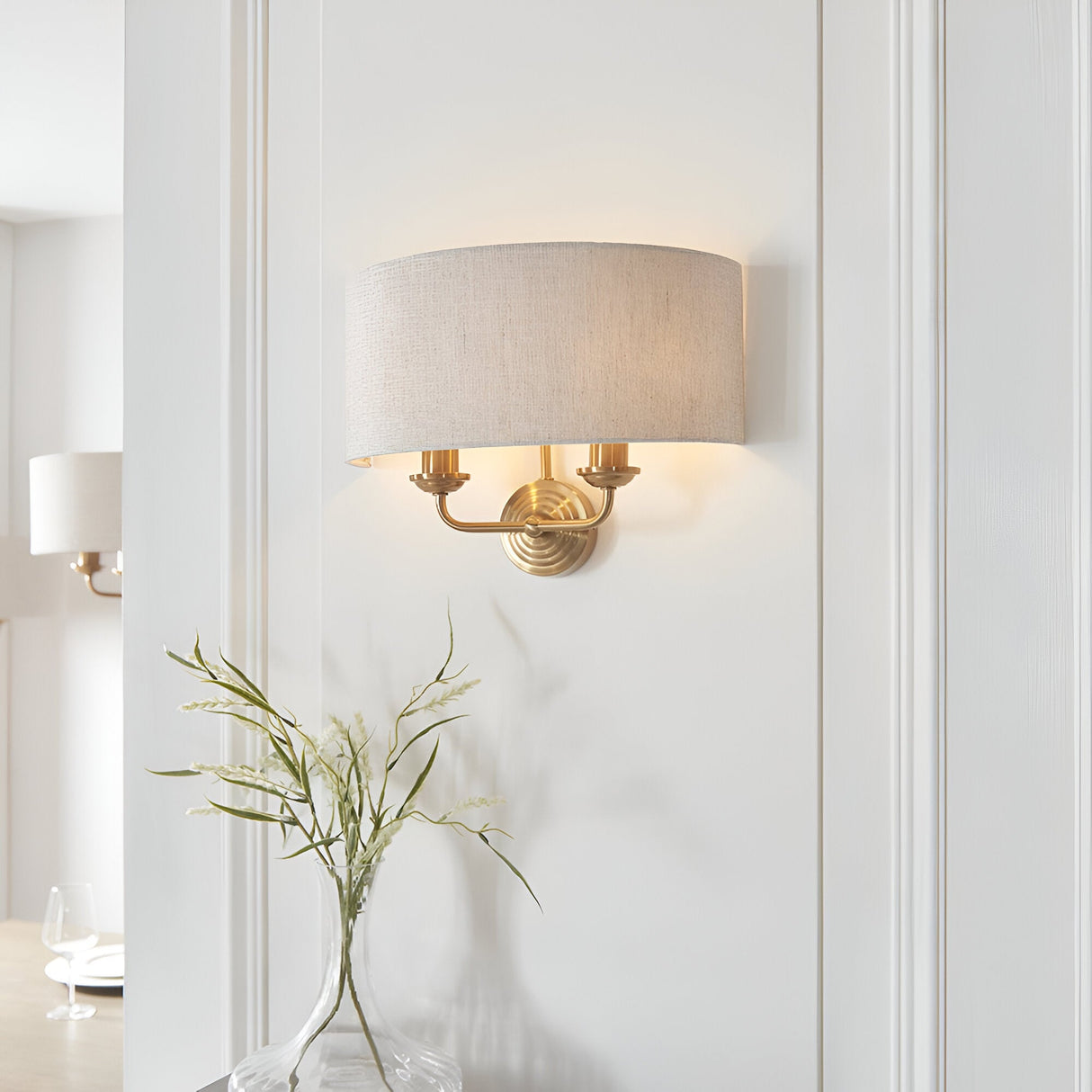 Endon Brenley 2 light wall light in vintage brass with natural linen drum shade, mounted on a white panelled wall above a glass vase of foliage.