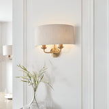 Endon Brenley 2 light wall light in vintage brass with natural linen drum shade, mounted on a white panelled wall above a glass vase of foliage.