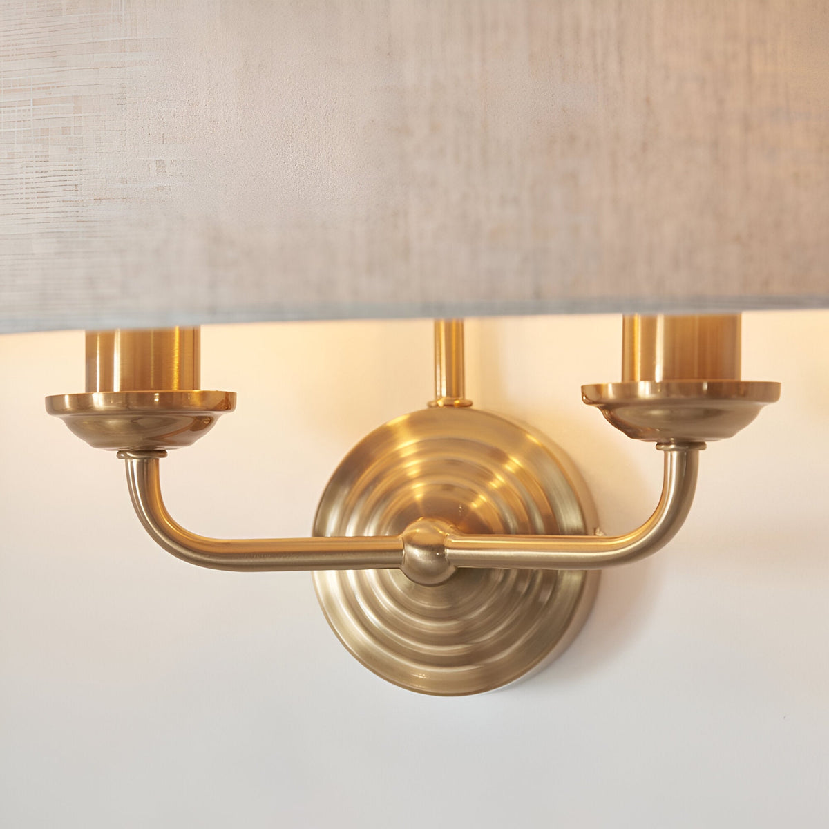Close-up of Endon Brenley 2 Light Wall Light in vintage brass, showing the round backplate, curved twin arms and base of a natural linen shade.