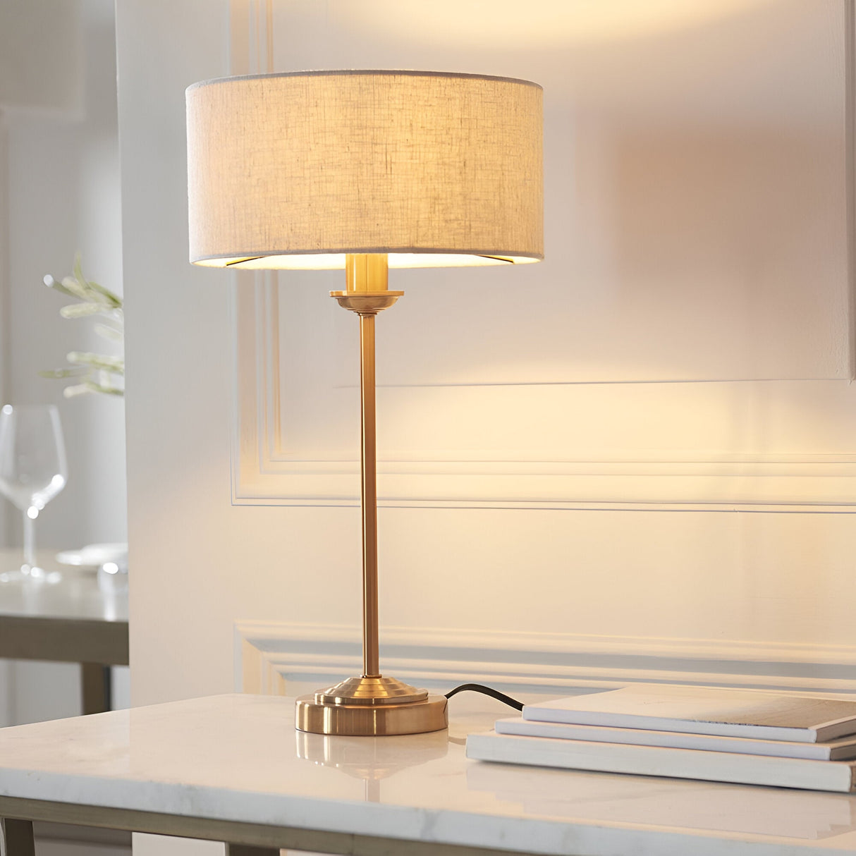 Endon Brenley brass table lamp with slim stem and natural linen drum shade, lit on a marble console table in a softly styled living space.