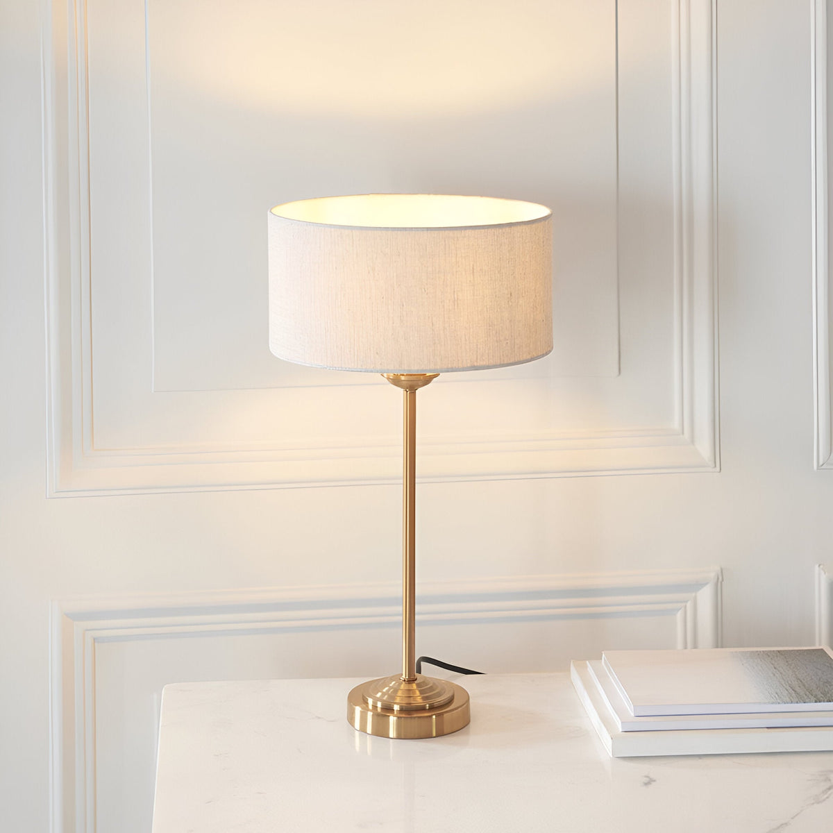 Endon Brenley table lamp in brass with a slim stem, stepped round base and natural linen drum shade, lit on a marble surface against panelled wall.