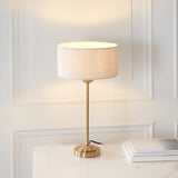 Endon Brenley table lamp in brass with a slim stem, stepped round base and natural linen drum shade, lit on a marble surface against panelled wall.
