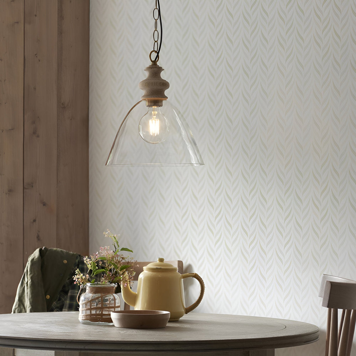 Bevwick pendant light with flared clear glass shade, ash wood top, and aged brass chain, shown above a dining table with lit E27 bulb.