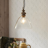 Bevwick pendant light with flared clear glass shade, turned ash wood top and black chain, shown lit above a dining table with wallpaper backdrop.