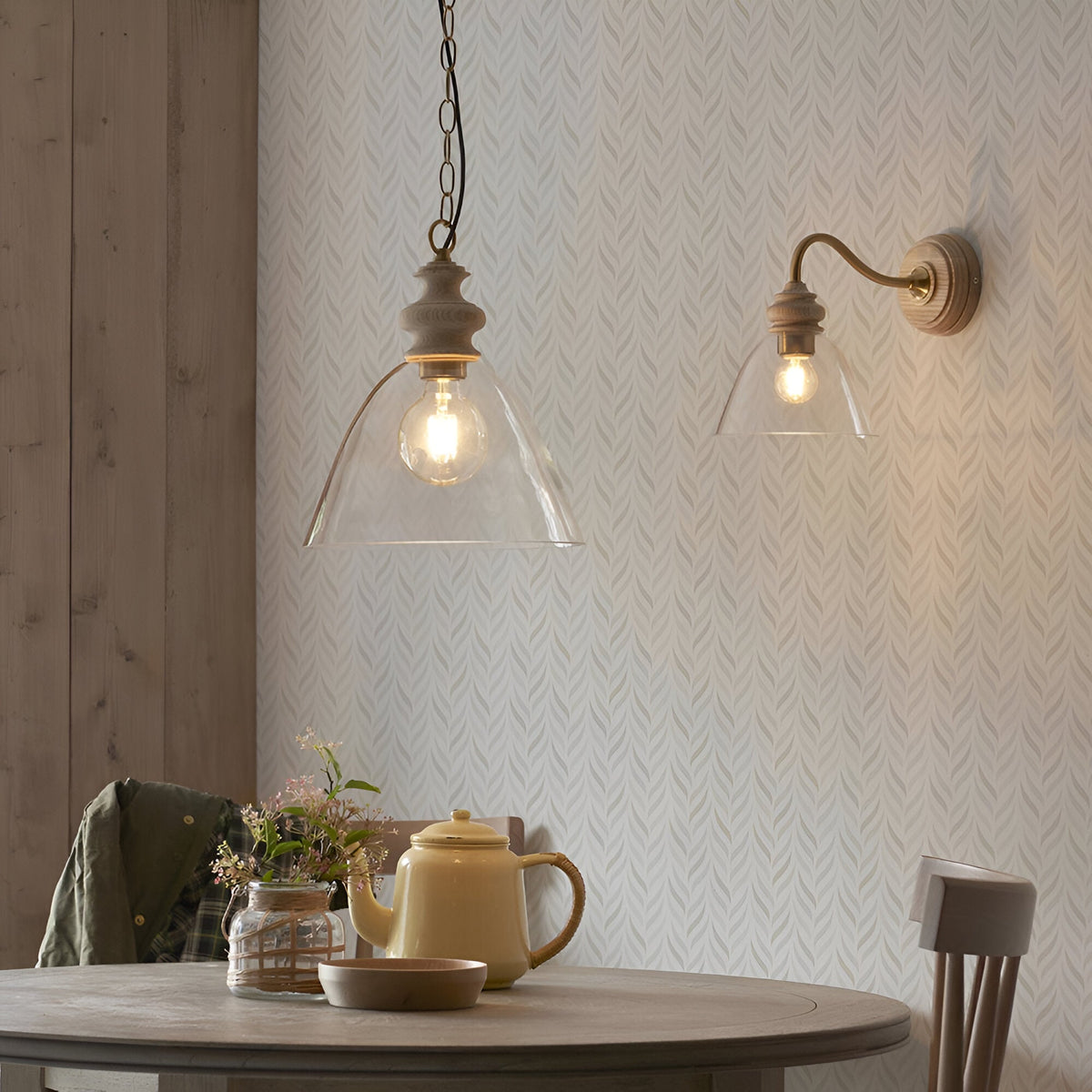 Bevwick Pendant with clear flared glass shade, ash wood top and aged brass chain, shown lit above a dining table beside a matching wall light.