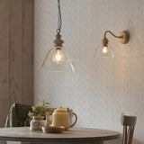 Bevwick Pendant with clear flared glass shade, ash wood top and aged brass chain, shown lit above a dining table beside a matching wall light.