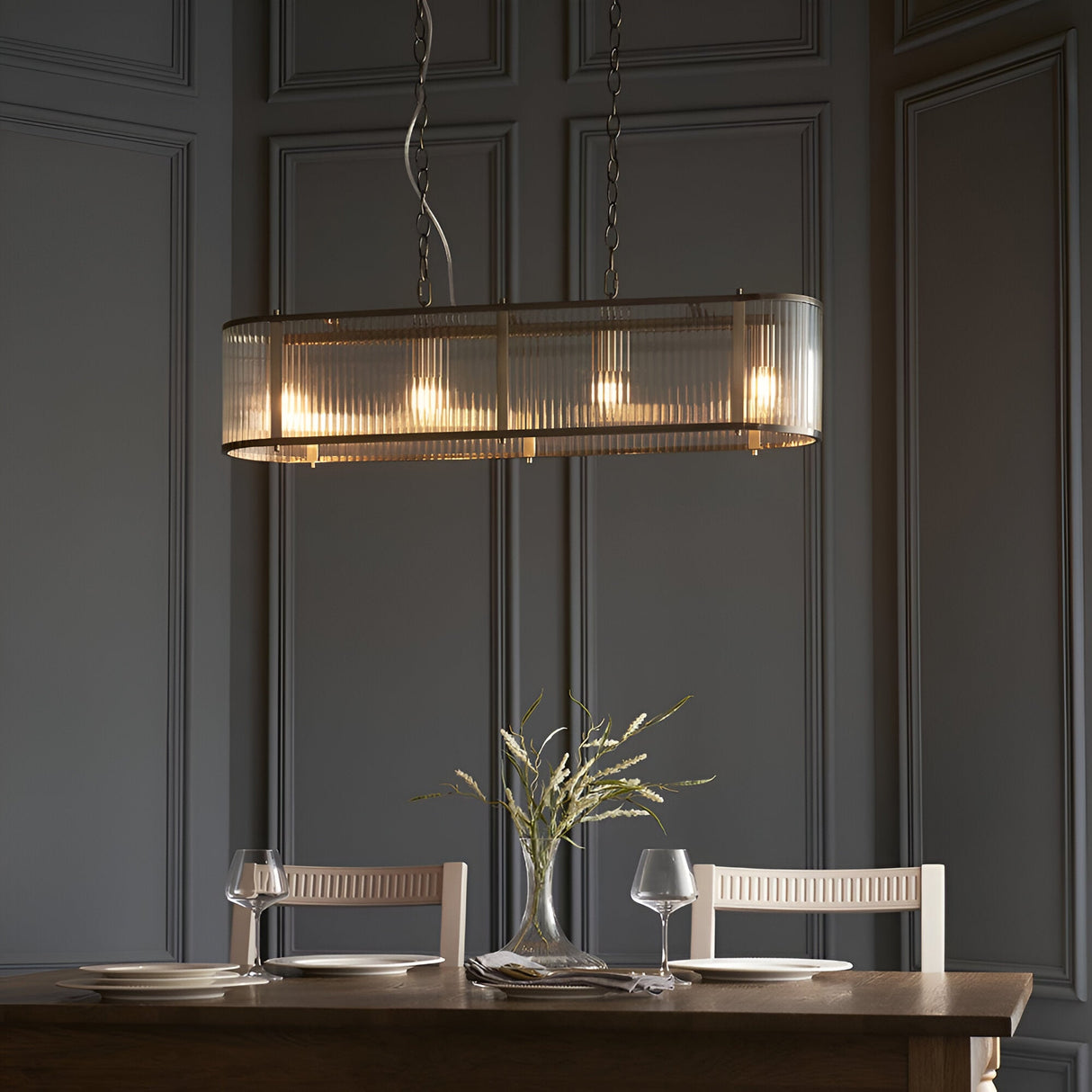 Rothbury 4 Light Linear Pendant in antique brass with ribbed glass shades, hung above a wood dining table in a dark panelled room.