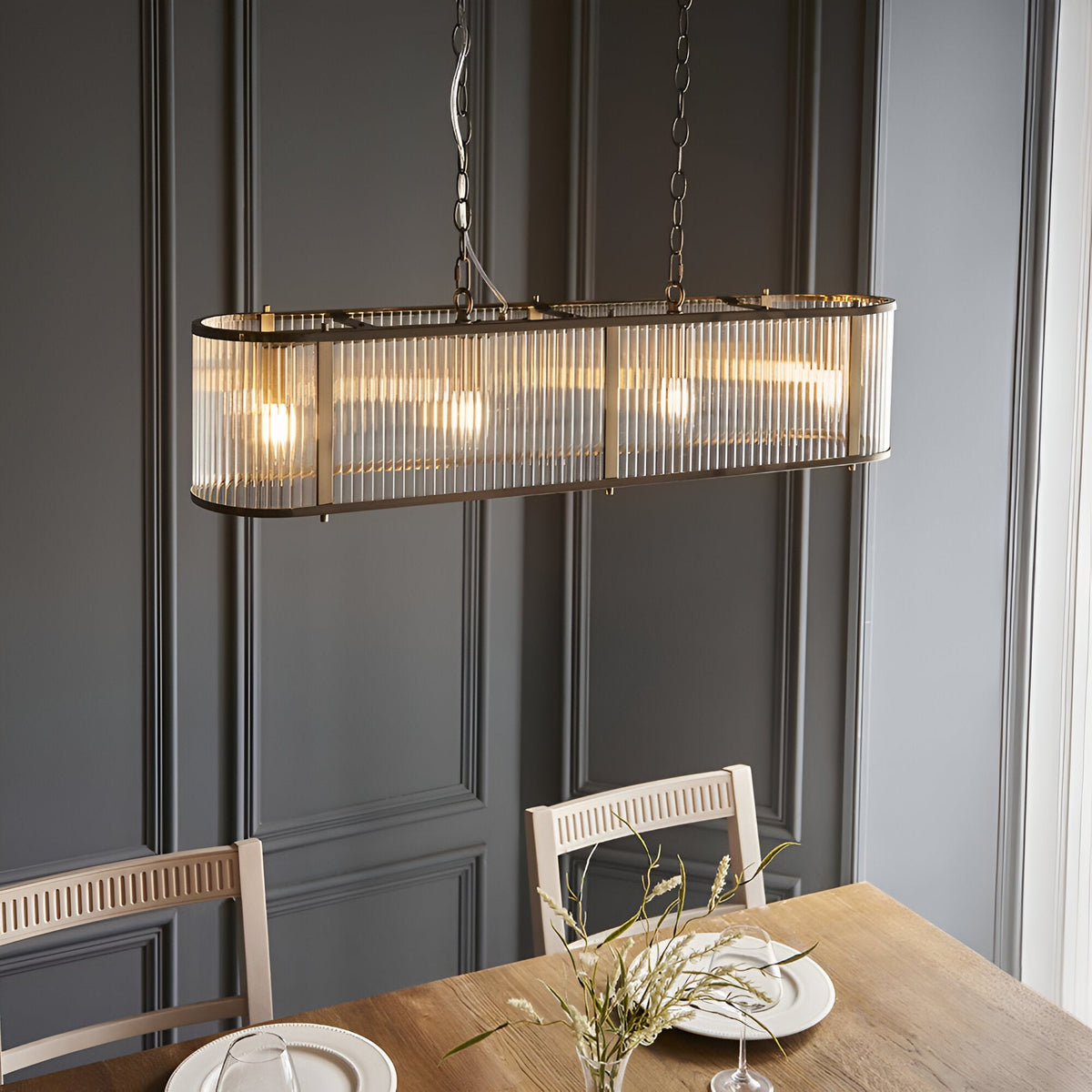 Rothbury 4 Light Linear Pendant in antique brass with ribbed glass shades, hung on chains above a dining table in a panelled dark grey room.