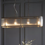 Rothbury 4 Light Linear Pendant in antique brass with ribbed glass shades, oval bar design, hung on chains against a dark panelled wall.