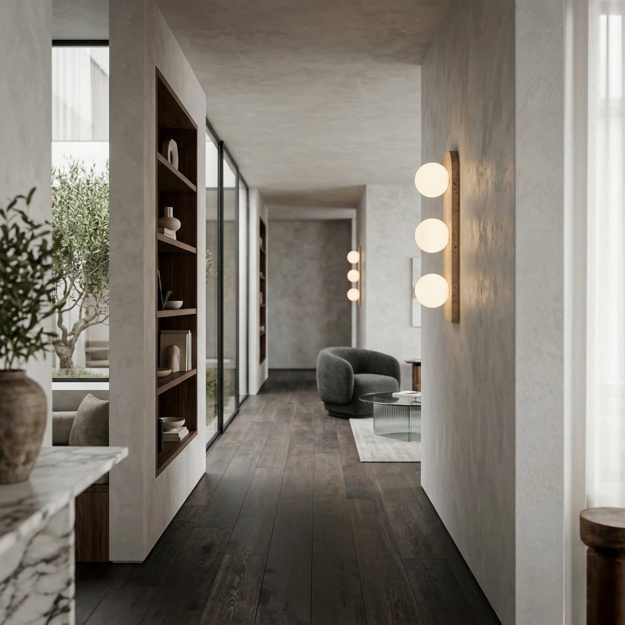 A modern hallway with dark wood floors, built-in shelves, large windows, and a cozy seating area featuring a gray chair and glass table is softly lit by Virella 3 Light Vertical Travertine Wall Lights in light beige; greenery is visible outside.