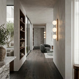 A modern hallway with dark wood floors, built-in shelves, large windows, and a cozy seating area featuring a gray chair and glass table is softly lit by Virella 3 Light Vertical Travertine Wall Lights in light beige; greenery is visible outside.