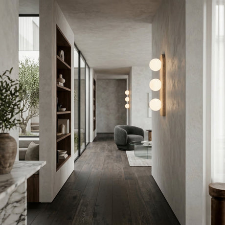 A modern hallway with dark wood floors, built-in shelves, large windows, and a cozy seating area featuring a gray chair and glass table is softly lit by Virella 3 Light Vertical Travertine Wall Lights in light beige; greenery is visible outside.