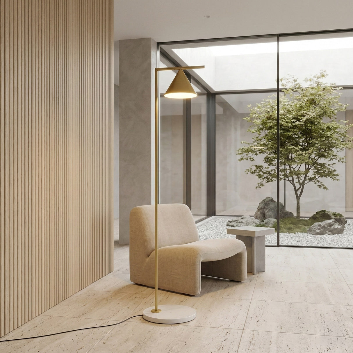 A modern minimalist room with a beige armchair, a small side table, and the Odin Floor Lamp - Brass. Large windows look out to a zen garden with a tree and rocks. Interior features include light wood paneling and a stone floor.