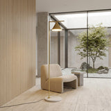 A modern minimalist room with a beige armchair, a small side table, and the Odin Floor Lamp - Brass. Large windows look out to a zen garden with a tree and rocks. Interior features include light wood paneling and a stone floor.
