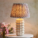 Amberley table lamp with a white bobble-textured ceramic base, aged brass stem, and pleated grey ikat shade, shown with pink flowers on a marble table.
