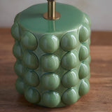 Close-up of the Endon Amberley ceramic table lamp base in glossy olive green with sculpted bobble texture and an aged brass stem, on a wooden surface.