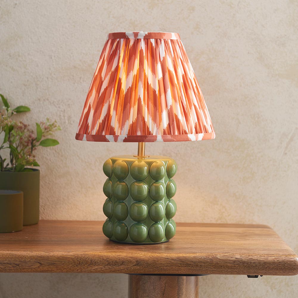 Endon Amberley table lamp with green ceramic bobble base, aged brass stem and pleated orange ikat-pattern fabric shade, on a wooden table.