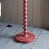 Endon Brenleigh floor lamp base in glossy pink with stacked metal spheres and a matching fabric flex, shown on grey plank flooring by a panelled wall.