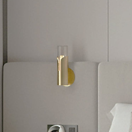 Bisel Cylinder Ribbed Glass Wall Light in Gold, mounted above a headboard, with a ribbed glass cylinder half-wrapped in a curved gold sleeve on a round backplate.