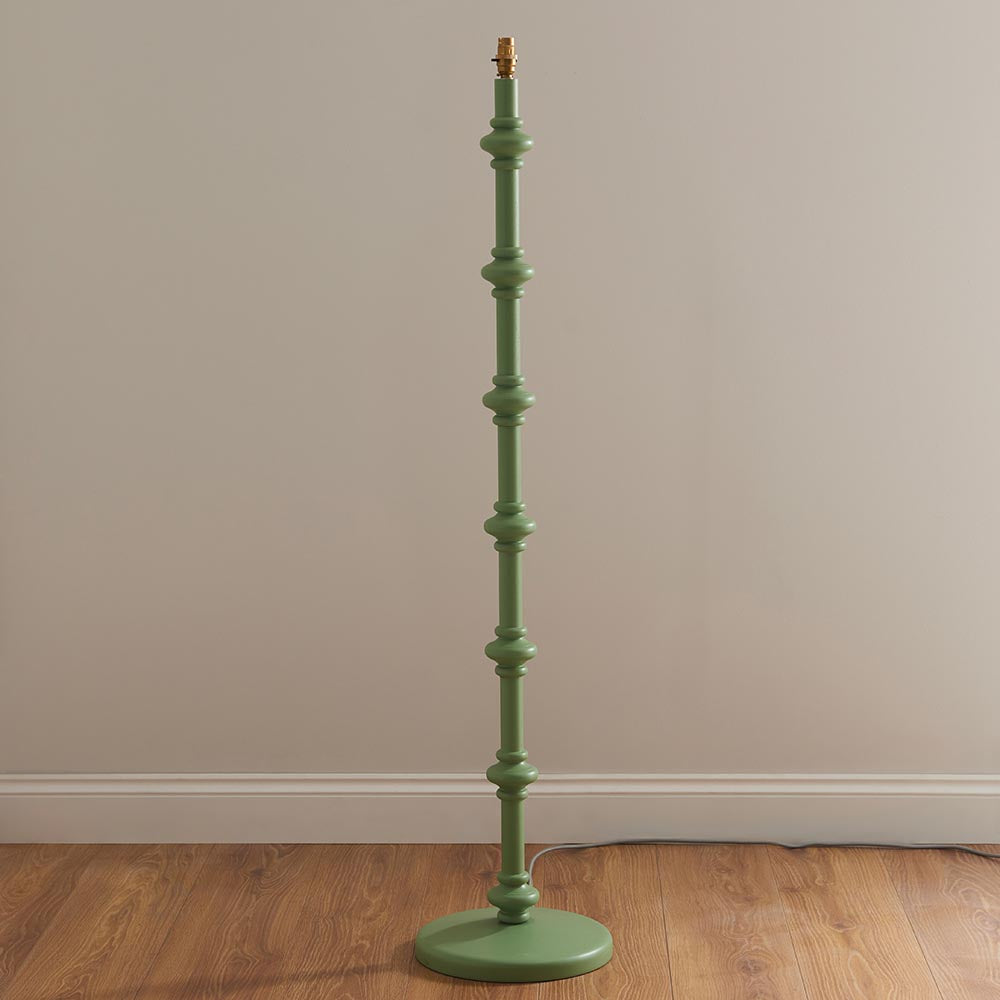Endon Bevbury floor lamp with a Cotswold green turned wooden spindle base and brass B22 lamp holder, shown against a neutral wall on wood flooring.