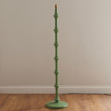 Endon Bevbury floor lamp with a Cotswold green turned wooden spindle base and brass B22 lamp holder, shown against a neutral wall on wood flooring.