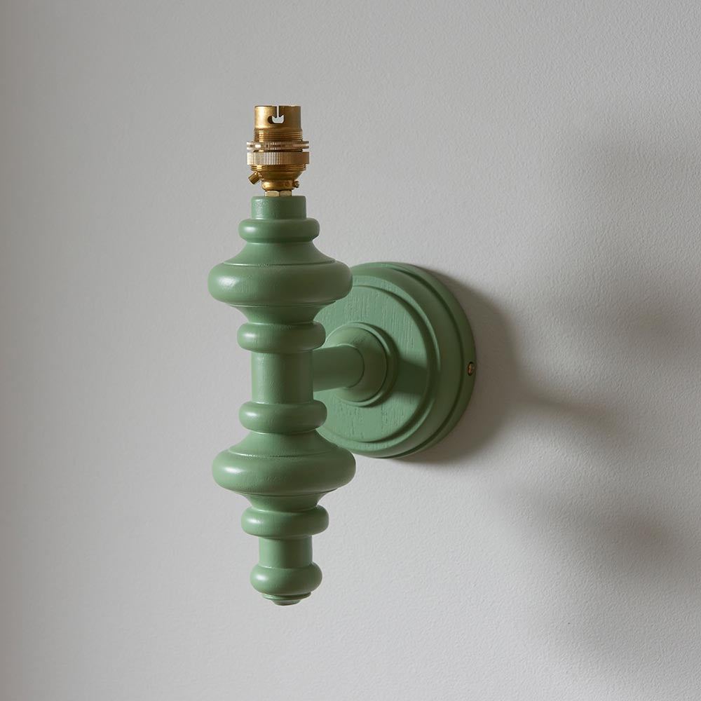 Endon Bevbury wall light with a turned wooden spindle in satin Cotswold green and an exposed brass B22 lamp holder, mounted on a light grey wall.