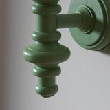 Close-up of the Endon Bevbury wall light showing its turned wooden spindle in Cotswold green satin finish, mounted on a round backplate against a pale wall.