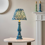 Endon Carlworth table lamp with gloss blue turned spindle base, brass holder and pleated blue patterned shade, on a marble side table by a fireplace.