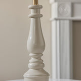 Carlworth table lamp base in white gloss resin with turned spindle design and brass B22 bayonet lamp holder, shown against a softly blurred interior background.